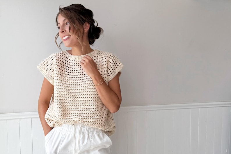 May include: Cream-colored, open-weave crochet top with short sleeves. The top has a round neckline and a loose fit. The model is wearing white pants. The background is a light gray wall.