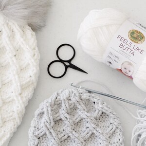 May include: White crochet hat with a fluffy pom pom, a skein of white yarn labeled "Feels Like Butta" Lion Brand Yarn, a pair of black scissors, a crochet hook, and a skein of gray yarn.