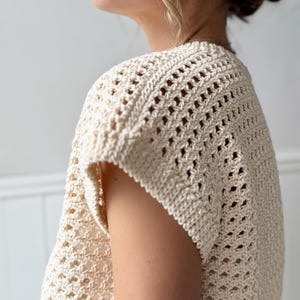May include: Cream-colored, open-weave crochet top. The short-sleeved garment features a textured, geometric pattern. The top is made of a soft, natural fiber, and the design allows for breathability. The style is casual and suitable for warm weather.