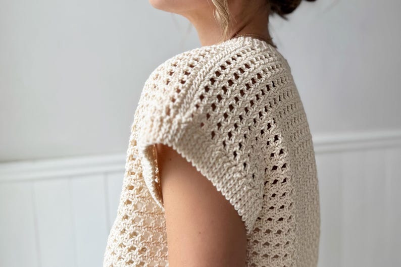 May include: Cream-colored, open-weave crochet top. The short-sleeved garment features a textured, geometric pattern. The top is made of a soft, natural fiber, and the design allows for breathability. The style is casual and suitable for warm weather.