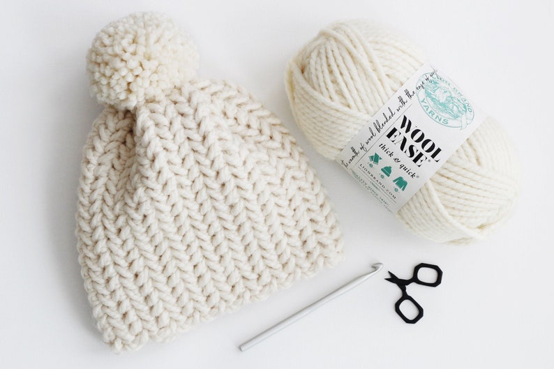 Pode incluir: Um gorro de tric&ocirc; branco com um pompom no topo, um novelo de l&atilde; branca, um gancho de croch&ecirc; e uma tesoura sobre uma superf&iacute;cie branca. O r&oacute;tulo da l&atilde; diz "Wool Ease Thick & Quick".