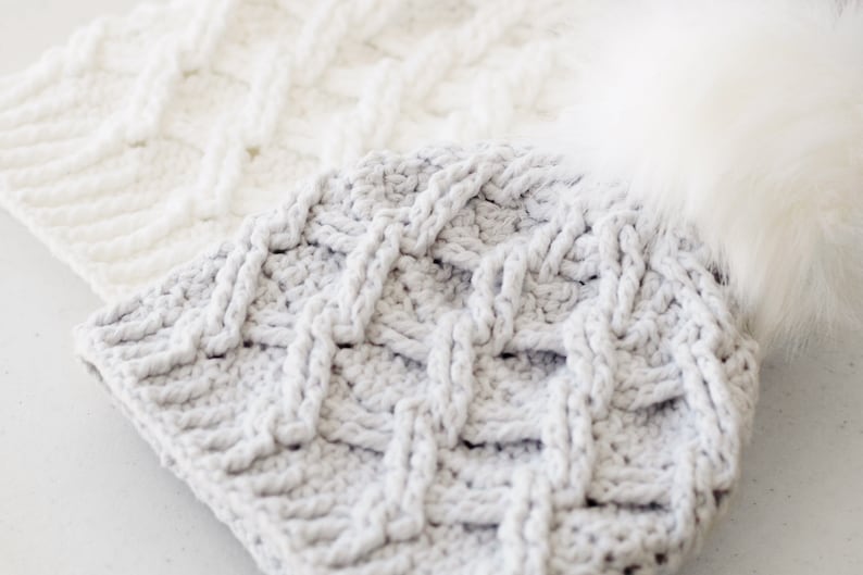 May include: Two crocheted hats, one white and one gray, with a braided pattern. The gray hat has a white faux fur pom pom.