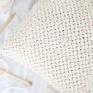 May include: A close-up of a white knitted pillow with a textured pattern. The pillow is made with a chunky yarn and has a soft, cozy look.