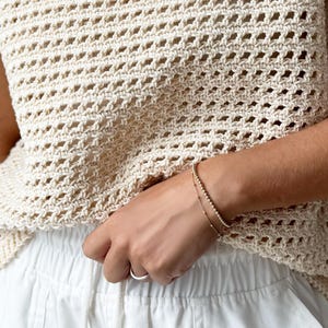 May include: A cream-colored, open-weave crochet top with a round neckline and short sleeves. The top is paired with white pants. A gold bracelet with small beads and a delicate chain bracelet are worn on the wrist.
