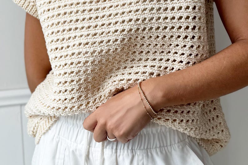 May include: A cream-colored, open-weave crochet top with a round neckline and short sleeves. The top is paired with white pants. A gold bracelet with small beads and a delicate chain bracelet are worn on the wrist.