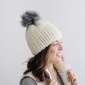 Pode incluir: Um gorro de croch&ecirc; branco com um pompom de pele sint&eacute;tica cinza. O gorro est&aacute; sendo usado por uma pessoa com cabelo castanho longo.