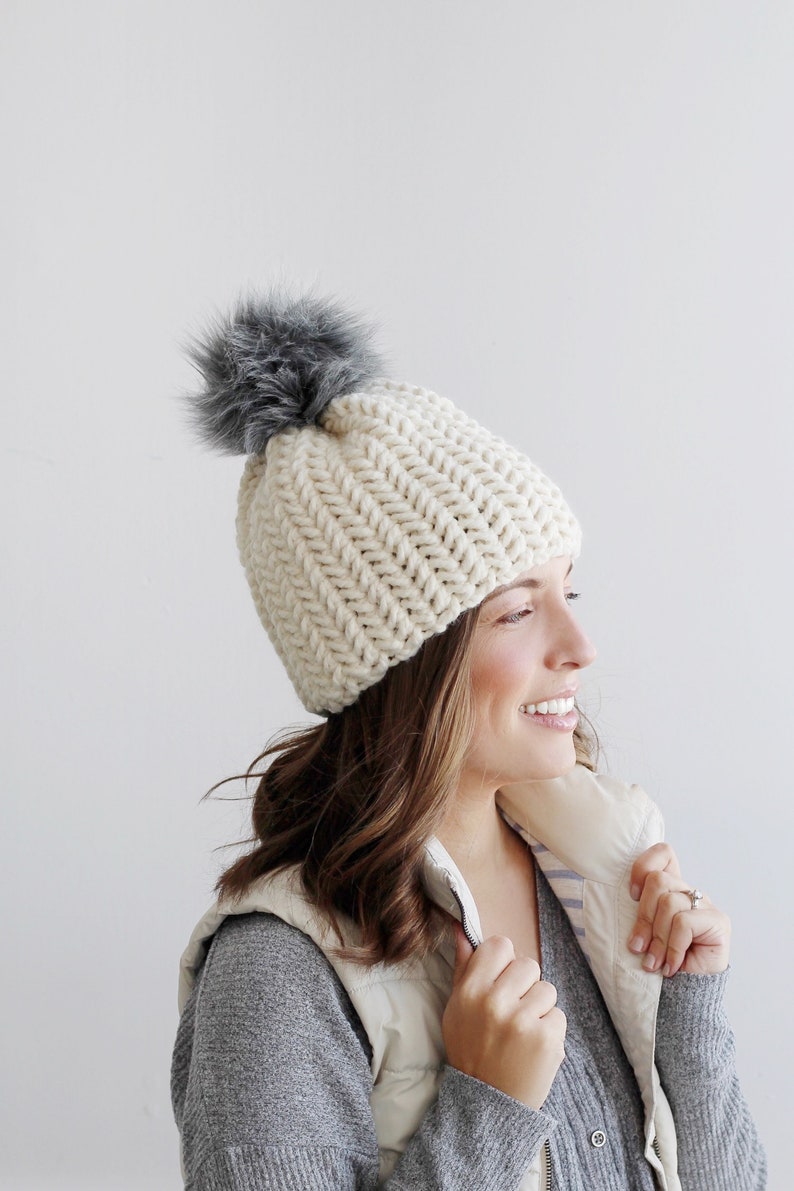 Pode incluir: Um gorro de croch&ecirc; branco com um pompom de pele sint&eacute;tica cinza. O gorro est&aacute; sendo usado por uma pessoa com cabelo castanho longo.