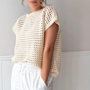 May include: A cream-colored, open-weave crochet top with short sleeves. The top has a loose, boxy fit and is paired with white pants. The crochet pattern creates a textured, airy look, suitable for warm weather. The top is a neutral color.
