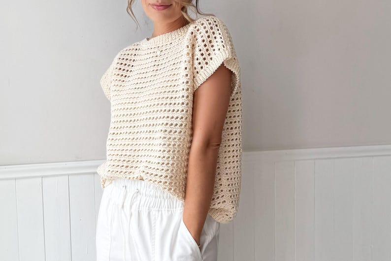 May include: A cream-colored, open-weave crochet top with short sleeves. The top has a loose, boxy fit and is paired with white pants. The crochet pattern creates a textured, airy look, suitable for warm weather. The top is a neutral color.