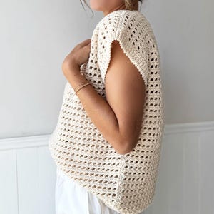 May include: Cream-colored, open-weave crochet top with short sleeves. The top has a loose, flowing fit and a textured appearance. The garment is made of a light-colored yarn and is suitable for warm weather.