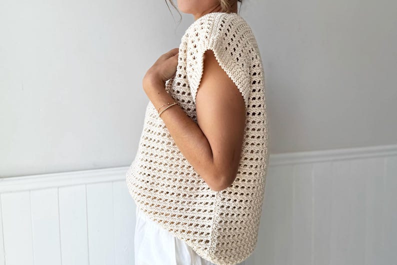 May include: Cream-colored, open-weave crochet top with short sleeves. The top has a loose, flowing fit and a textured appearance. The garment is made of a light-colored yarn and is suitable for warm weather.