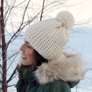 Pode incluir: Um gorro de malha branco com um pompom no topo. O gorro tem um padr&atilde;o de ponto espinha de peixe. O gorro &eacute; usado por uma pessoa que usa uma jaqueta verde com capuz forrado de pele.