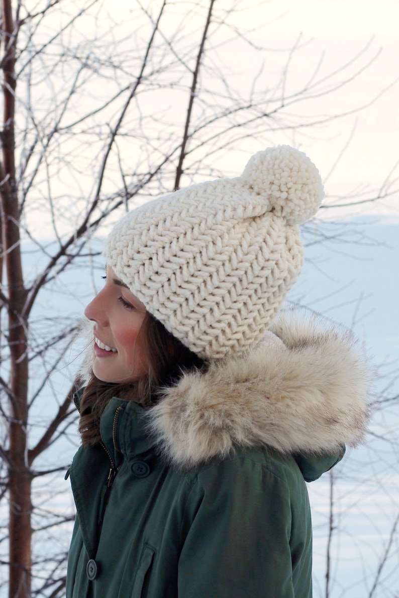 Pode incluir: Um gorro de malha branco com um pompom no topo. O gorro tem um padr&atilde;o de ponto espinha de peixe. O gorro &eacute; usado por uma pessoa que usa uma jaqueta verde com capuz forrado de pele.