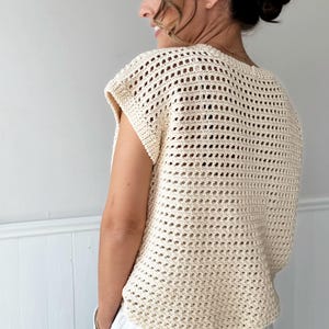 May include: Cream-colored crochet top with a loose, open weave design. The top has a round neckline and short sleeves. The model is wearing white pants. The top is a light, airy garment, perfect for warm weather.