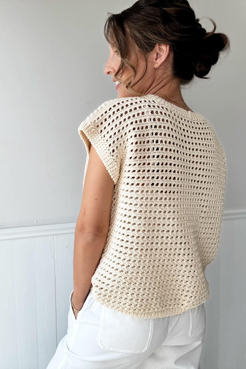 May include: Cream-colored crochet top with a loose, open weave design. The top has a round neckline and short sleeves. The model is wearing white pants. The top is a light, airy garment, perfect for warm weather.
