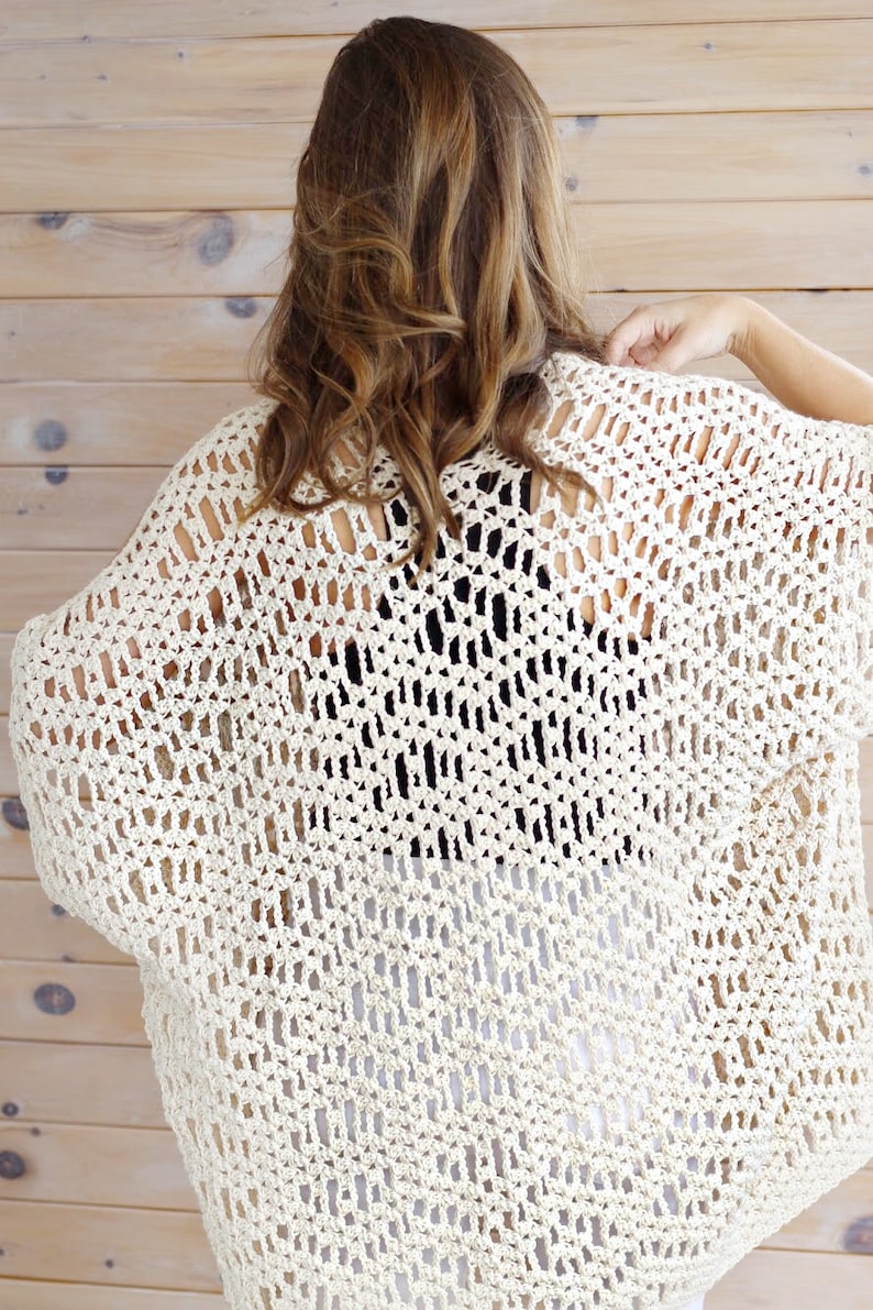 May include: A crocheted cream-colored cardigan with a loose, open-weave design. The cardigan is shown from the back and has a slightly cropped length.