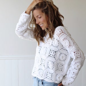 May include: White crochet sweater with a square pattern and pom-poms. The sweater has long sleeves and a relaxed fit.