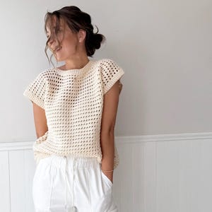 May include: Cream-colored crochet top with a loose, open weave design. The top has short sleeves and a round neckline. Paired with white pants. The outfit is casual and summery.