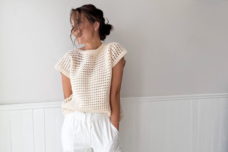 May include: Cream-colored crochet top with a loose, open weave design. The top has short sleeves and a round neckline. Paired with white pants. The outfit is casual and summery.