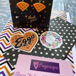May include: Waffle-shaped earrings with silver hooks are displayed in front of a black gift bag with white, orange, and purple polka dots. A sticker with the word "Eggo" on a waffle shape and a floral sticker are also visible.