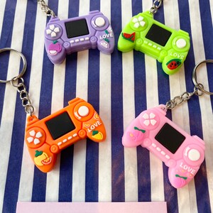 May include: Four brightly coloured video game controller keyrings. The controllers are purple, green, orange, and pink, each with a black screen and fruit-themed decorations. Each keyring has a silver ring and chain. The word "Love" is on each controller.