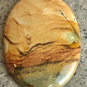 Puede incluir: Una piedra pulida de forma ovalada con un patrón de remolino de colores marrón, amarillo y verde. La piedra tiene un acabado liso y brillante.