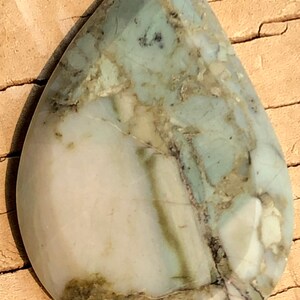 Puede incluir: Un cabujón en forma de lágrima de piedra con patrones verdes y blancos. La piedra tiene un aspecto moteado con remolinos de verde y blanco.