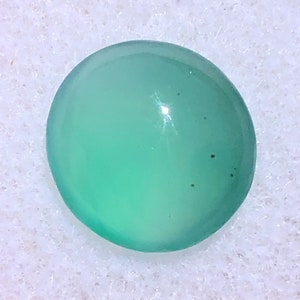 Può includere: Una pietra preziosa rotonda di colore verde chiaro con una superficie liscia e lucida. La pietra presenta alcune piccole inclusionesioni scure.