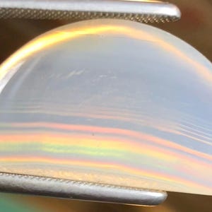 May include: A white agate cabochon with iridescent rainbow banding. The cabochon is shaped like a half-dome and is held in a pair of tweezers.