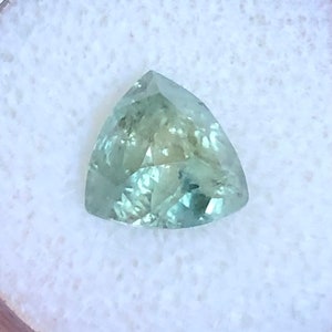 May include: A triangular cut green gemstone with a light green hue. The gemstone is set in a white background.