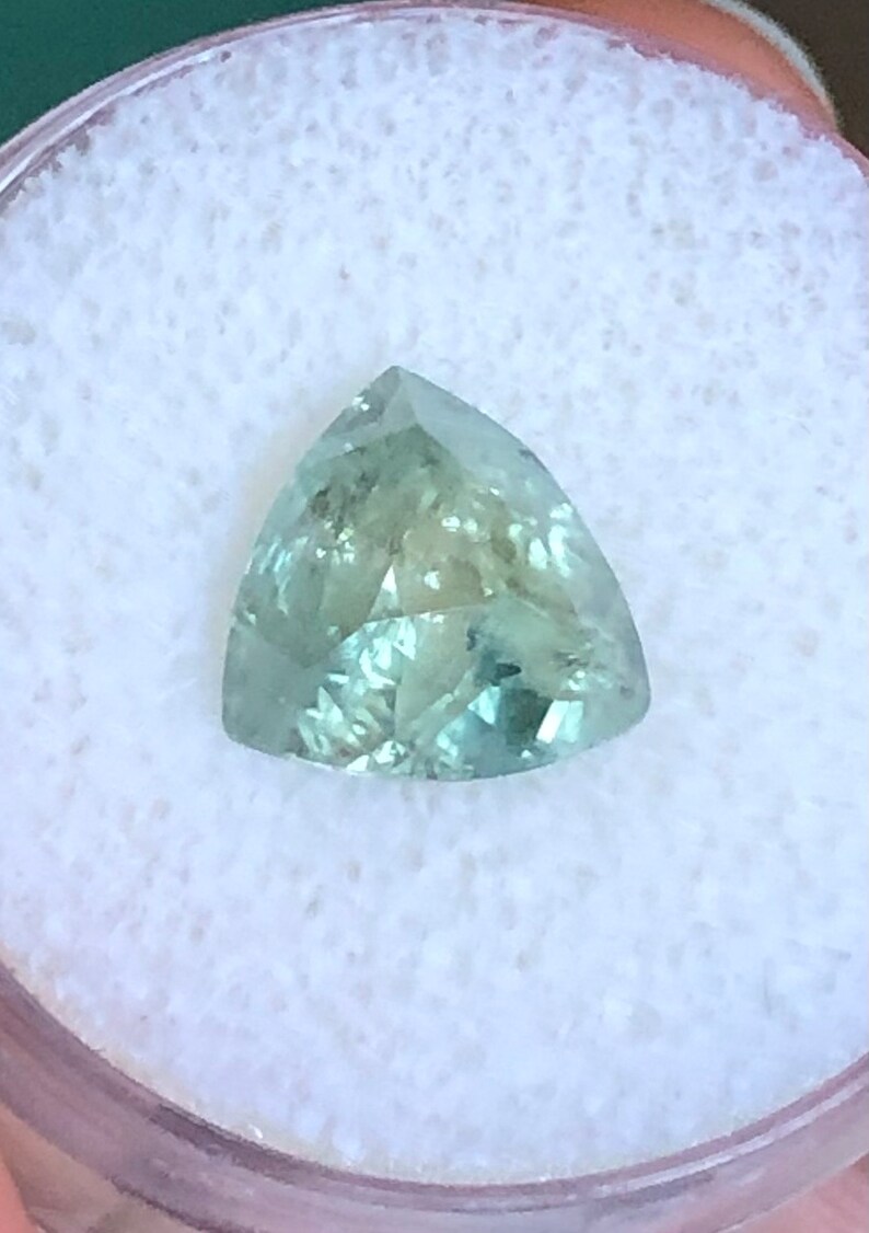 May include: A triangular cut green gemstone with a light green hue. The gemstone is set in a white background.