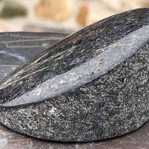 May include: A polished, dark gray stone object with a rounded, angled shape. The stone has a speckled texture and a smooth, reflective surface on the top edge. The object appears to be a decorative piece or a paperweight.
