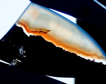 May include: A polished agate cabochon featuring a black, white and orange colour pattern. The agate has a smooth, rounded surface and a natural, organic shape.