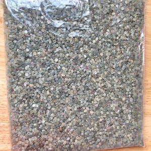 Puede incluir: Una bolsa de plástico transparente llena de pequeñas piedras preciosas rugosas de color gris, verde y rosa. La bolsa está sobre una superficie de madera.