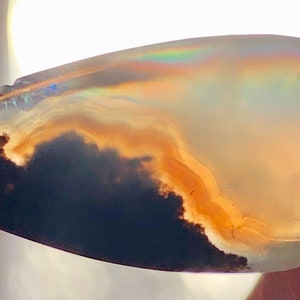 Puede incluir: Un cabujón de ágata en forma de lágrima con un patrón nublado, marrón y negro. La piedra tiene una calidad translúcida e iridiscente.