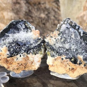 Könnte beinhalten: Zwei rohe Mineralproben in Grau, Schwarz und Weiß mit einer Matrix in Braun, Beige und Weiß. Die Proben befinden sich auf einem durchsichtigen Ausstellungsständer.