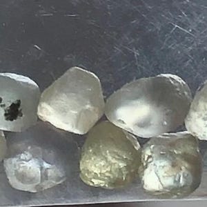 May include: A selection of rough gemstones in shades of pale yellow, green, and clear. The uncut stones have irregular shapes, appearing as raw, natural crystals. The image is taken in a clear plastic bag.