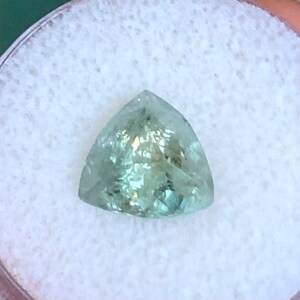May include: A triangular, faceted gemstone with a light green color. The gemstone is set in a white, circular container.