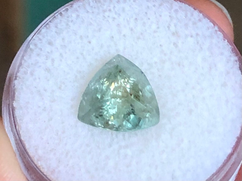 May include: A triangular, faceted gemstone with a light green color. The gemstone is set in a white, circular container.