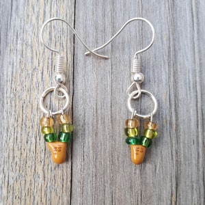 Pode incluir: Um par de brincos de prata com contas verdes e douradas e um amuleto de cerâmica marrom com o texto "105 + 35" em cada brinco.