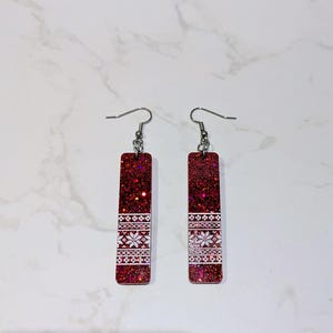 May include: A pair of rectangular drop earrings featuring a deep red glitter finish. The lower portion of each earring is decorated with a white, intricate, repeating geometric snowflake pattern, suspended from silver-toned metal earring hooks.