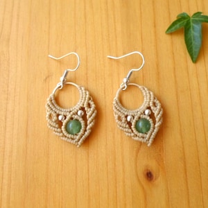 May include: A pair of beige macrame earrings with green stone accents. The earrings have a circular design with a teardrop shape at the bottom.