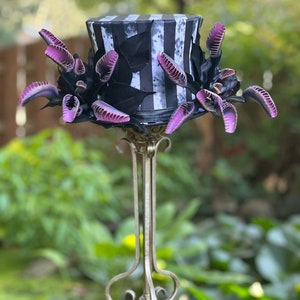 Puede incluir: Un sombrero de copa a rayas blancas y negras con un diseño de hojas blancas y negras. El sombrero está decorado con adornos florales de color morado y negro. El sombrero está sentado en un soporte de metal plateado.