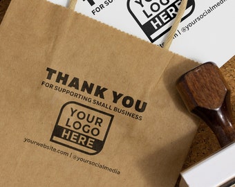 May include: A brown paper bag with a twisted paper handle and the text "THANK YOU FOR SUPPORTING SMALL BUSINESS" printed in black. A white card with the same text and a logo template is visible. A wooden stamp is also in the image.