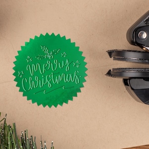May include: A green foil seal with the words "Merry Christmas" on a kraft paper envelope. A black embossing tool is positioned next to the seal. A pine cone and evergreen sprig are also in the image.