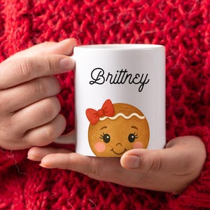 May include: White ceramic mug featuring the name "Brittney" in black script, above a gingerbread cookie design with a red bow. The mug is held against a red knitted sweater.