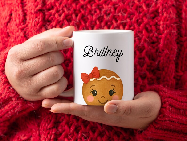 May include: White ceramic mug featuring the name "Brittney" in black script, above a gingerbread cookie design with a red bow. The mug is held against a red knitted sweater.
