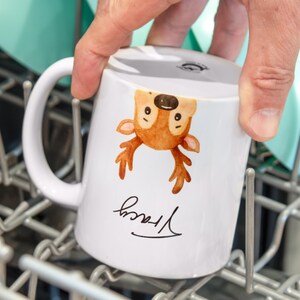 May include: White ceramic mug featuring a watercolor illustration of a deer with brown antlers and a smiling face. The name "Tracy" is written in cursive below the deer. The mug is being held in a dishwasher.