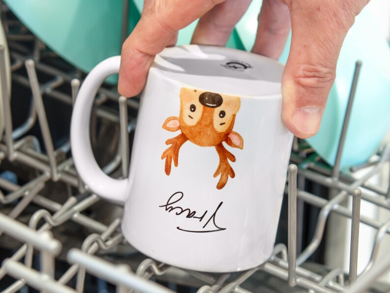 May include: White ceramic mug featuring a watercolor illustration of a deer with brown antlers and a smiling face. The name "Tracy" is written in cursive below the deer. The mug is being held in a dishwasher.