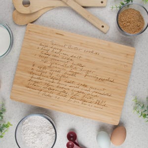 Large rectangular bamboo board shown with a "Peanut Butter Cookies" recipe engraved on it. A few kitchen items such as flour, milk, brown sugar, measuring tools, & spoons are displayed around the cutting board